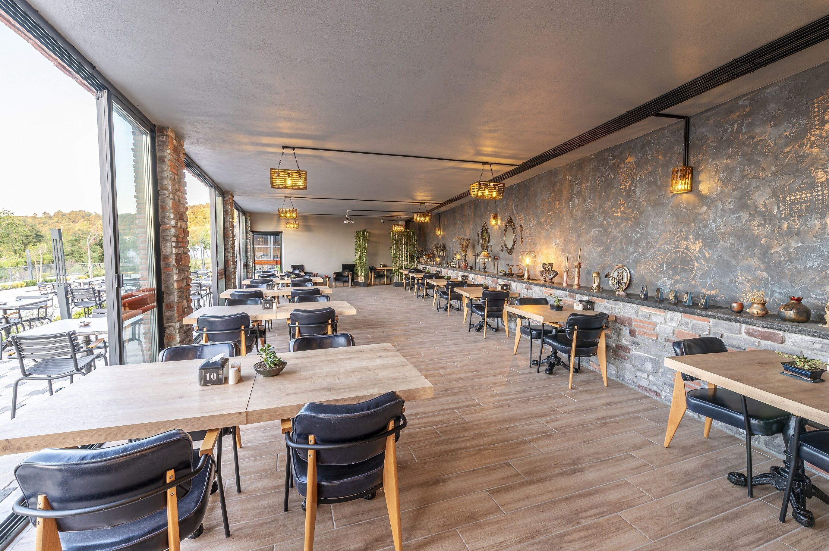 Un restaurante amplio con mesas y sillas largas, pared de ladrillo con adornos y ventanas grandes que ofrecen vistas al exterior.