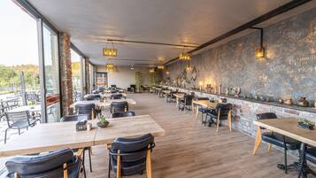 Un restaurante amplio con mesas y sillas largas, pared de ladrillo con adornos y ventanas grandes que ofrecen vistas al exterior.