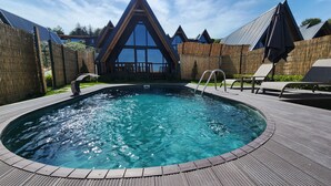Una piscina amb un tramat de fusta, gandules i un edifici al fons amb teulades triangulars.