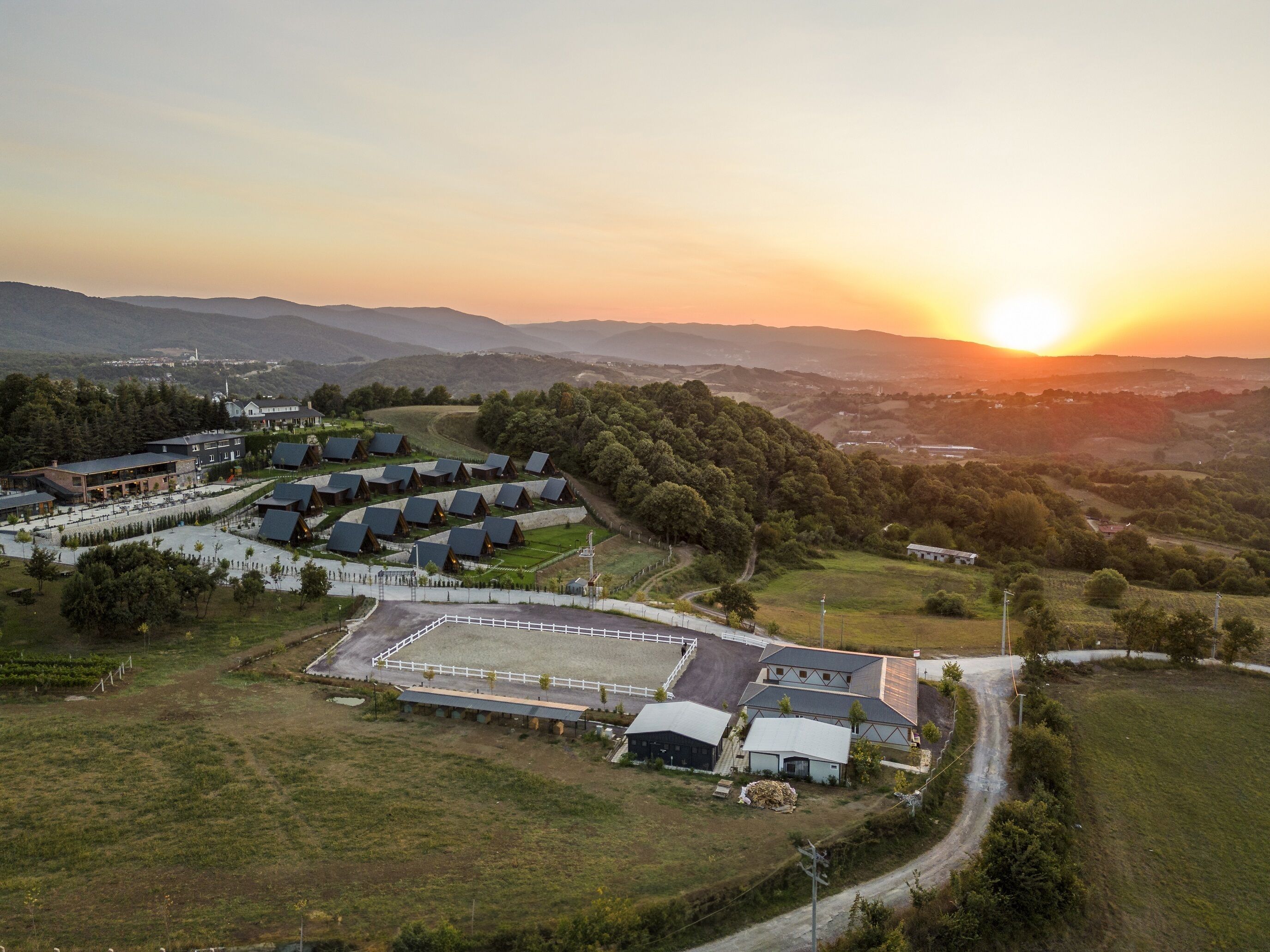 A rural setting with a large open field, several buildings with sloped roofs, and a sunset in the background.