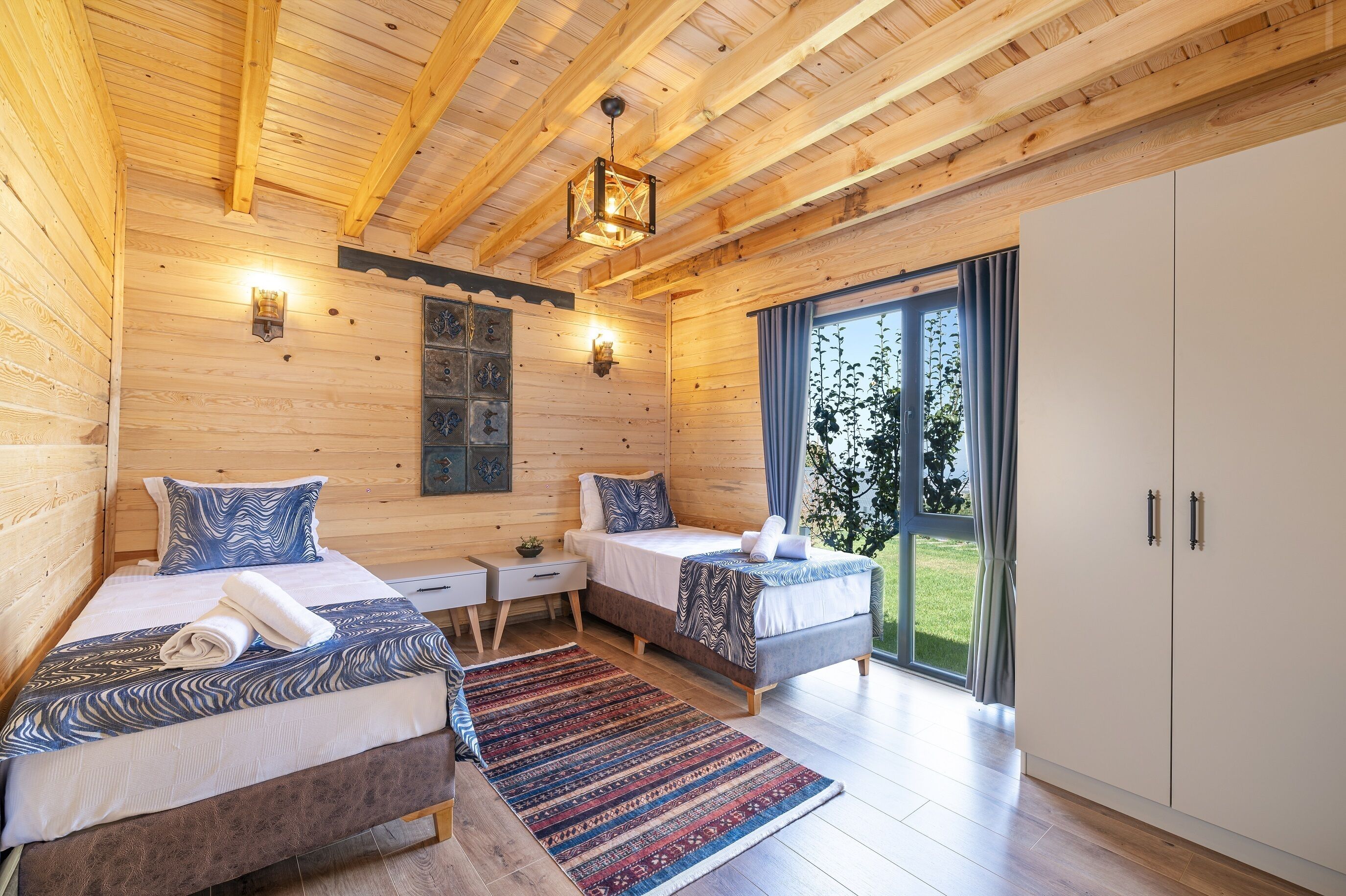 A wooden cabin room with two beds, a large window, and a wardrobe.
