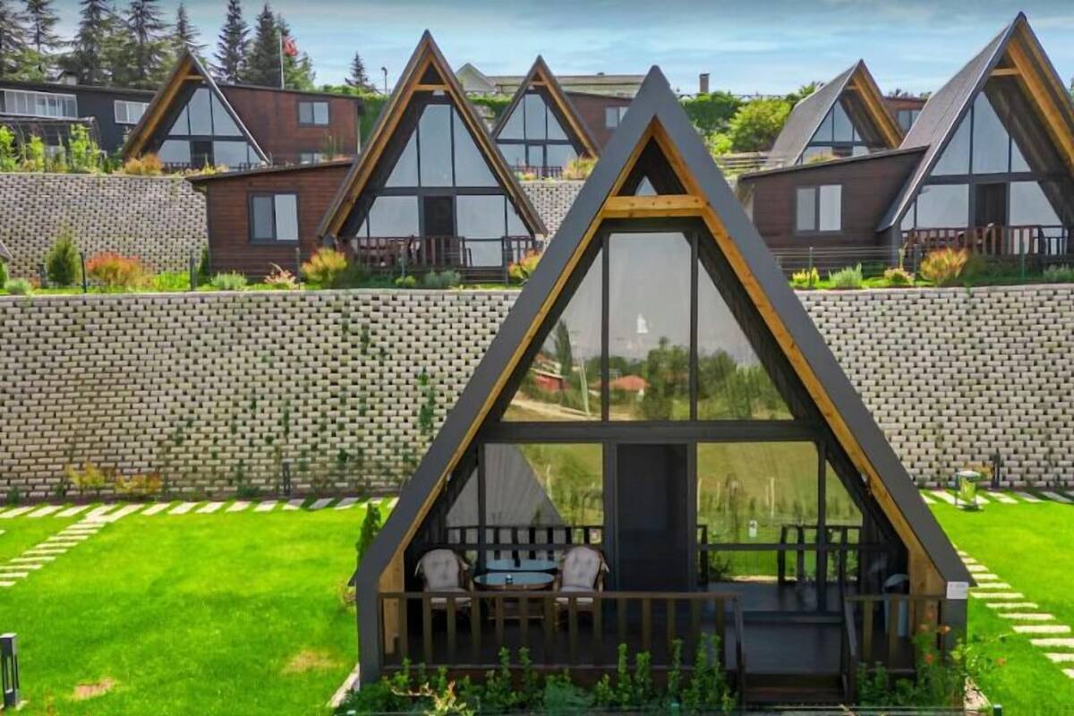 Una casa moderna de forma triangular con fachada de vidrio, un pequeño porche con mobiliario y un jardín bien cuidado.