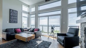 A spacious living room with a large window overlooking a body of water, a black leather sofa, a wooden coffee table, and a black armchair. - Hôtel à la maison - Entre brise et marée (Petite-Rivière-Saint-François)