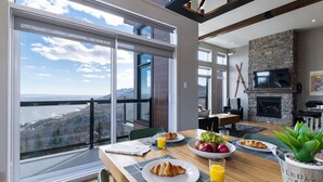 A dining area with a large table set for breakfast, a stone fireplace, and a scenic view through large windows. - Hôtel à la maison - Entre brise et marée (Petite-Rivière-Saint-François)