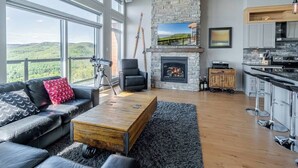 A modern living room with a stone fireplace, a leather sofa, and a large window overlooking a scenic landscape. - Hôtel à la maison - Entre brise et marée (Petite-Rivière-Saint-François)