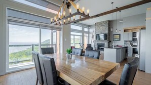 A modern kitchen with a stone fireplace, large windows, and a dining area with a wooden table and chairs. - Hôtel à la maison - Entre brise et marée (Petite-Rivière-Saint-François)