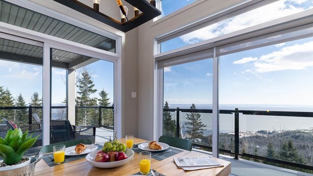 A breakfast table with croissants, fruit, and juice, overlooking a scenic view of trees and water. - Hôtel à la maison - Entre brise et marée (Petite-Rivière-Saint-François)