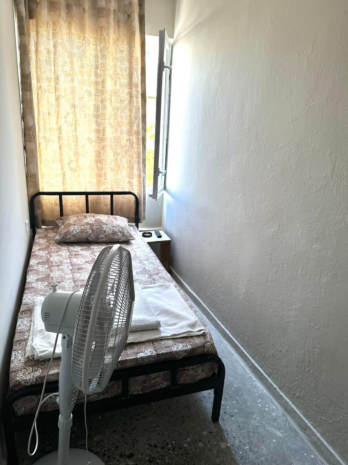A small, narrow room with a single bed, a standing fan, and a window with curtains.