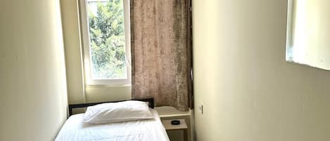 Quarto de hotel pequeno e estreito com uma cama de solteiro, mesa de cabeceira e janela com cortinas. Há também um ventilador fixado na parede.