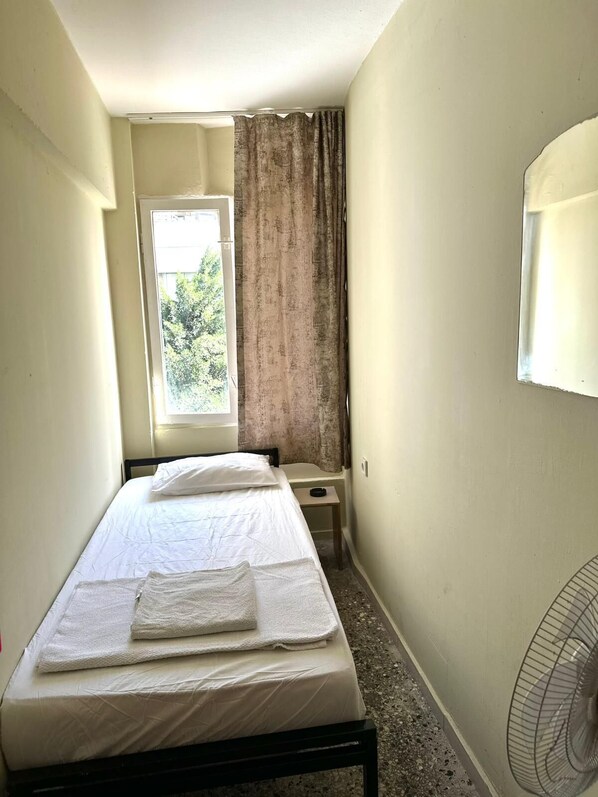 A narrow hotel room with a single bed, a small bedside table, a window with curtains, and a wall-mounted fan.