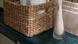 Cesto di vimini con asciugamani bianchi su una superficie scura, vicino a un vaso con piume bianche.