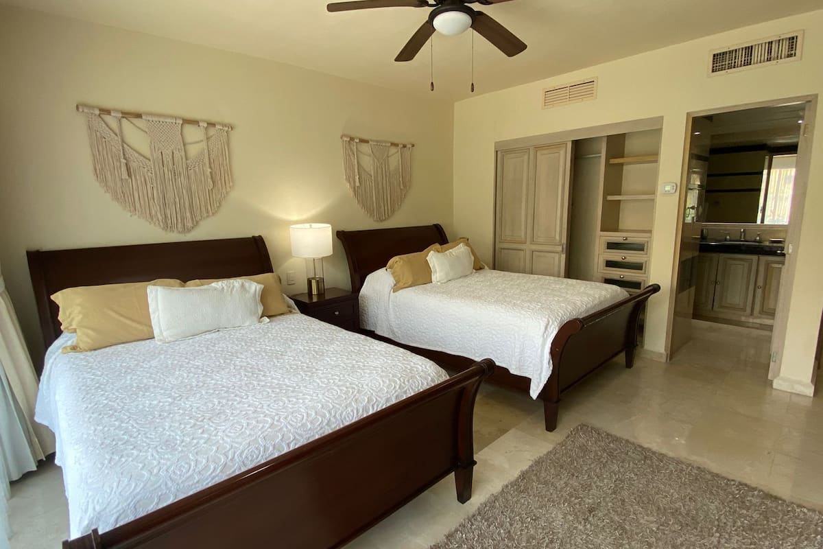A hotel room with two beds, a ceiling fan, a nightstand with a lamp, and a view of the kitchen.