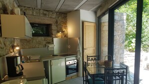 A compact kitchen with a stone wall, a wooden door, and a dining area with a glass table and chairs. - Cevennes Nature Entourée de Végétation, à 3mn de la Rivière, à 1mn de la Piscine (Chambon)