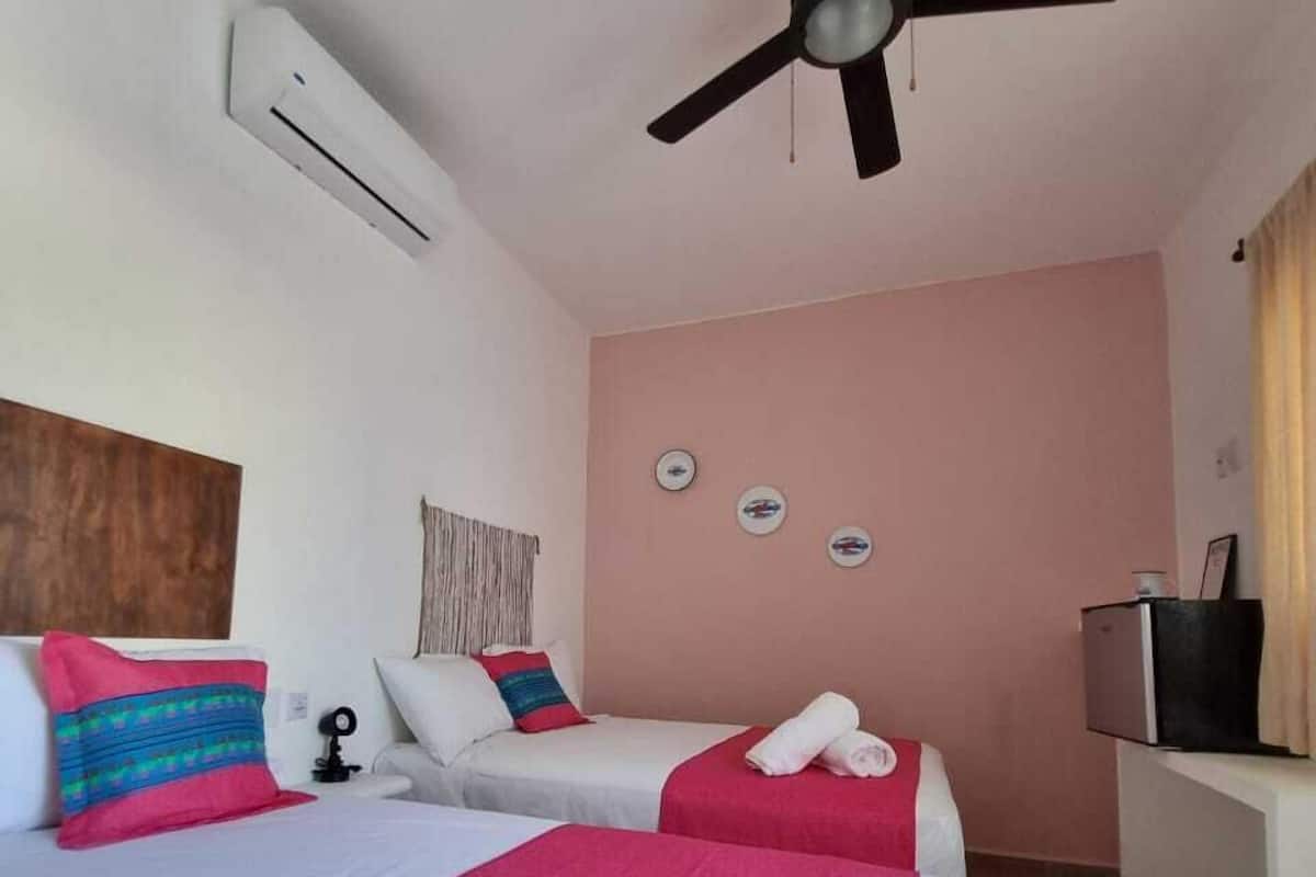 Ein Hotelzimmer mit zwei Betten, einem Deckenventilator, einer Klimaanlage, einem Fernseher und einem kleinen Schreibtisch mit Lampe.