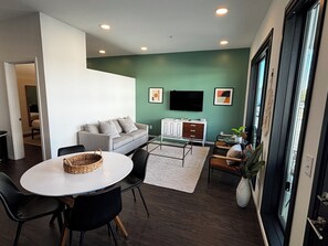 A modern living room with a sofa, a TV, a dining table with chairs, and a large window.