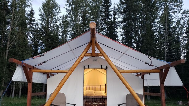 A wooden tent with a bed and two chairs, set on a deck with a forest backdrop. - Glamping tent close to the river by Sundre, Alberta (James River Bridge)