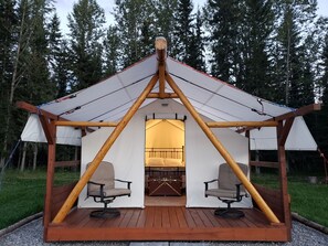 Une tente en bois équipée d’un lit et de deux chaises, installée sur une terrasse avec une forêt en arrière-plan.