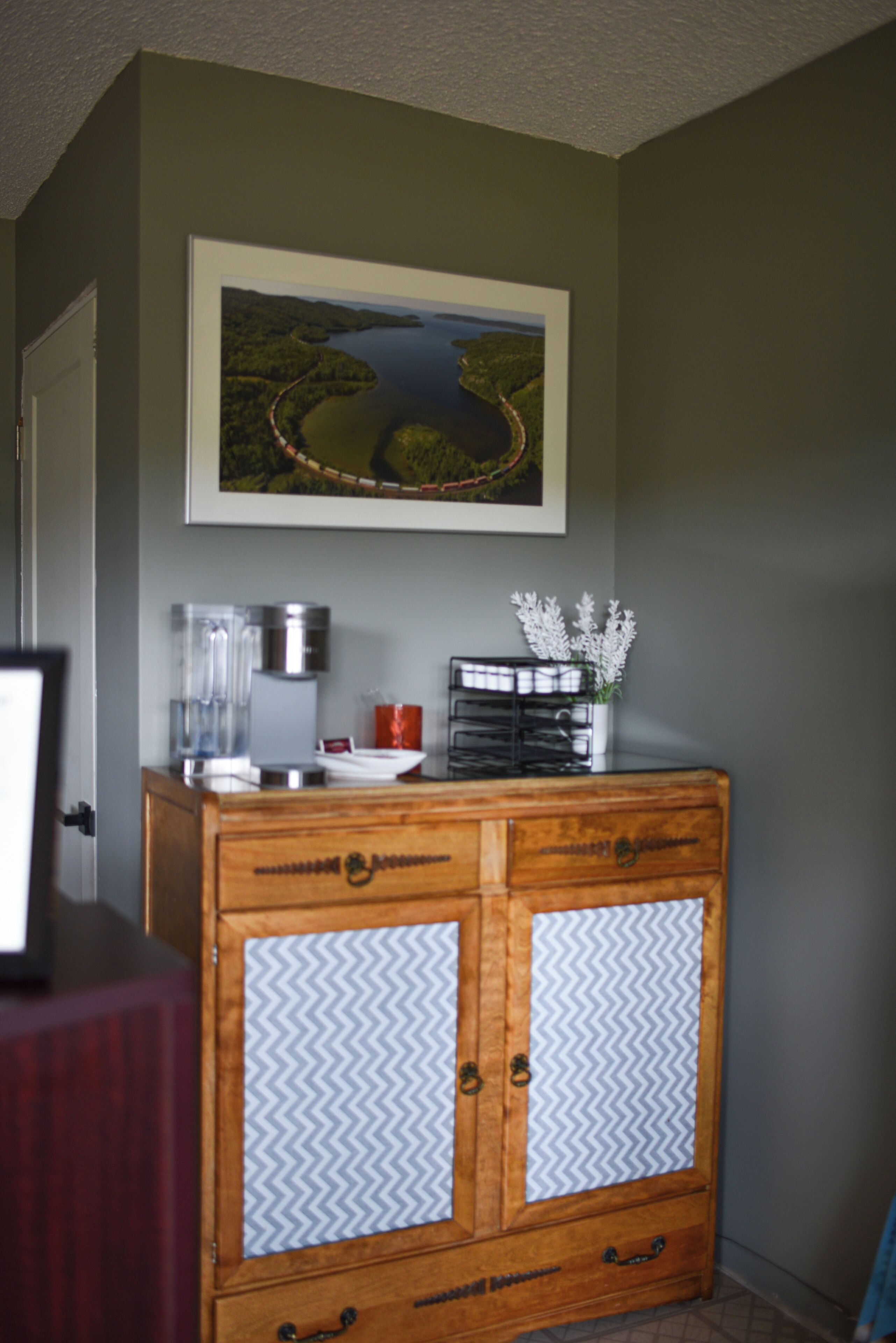 A room with a wooden cabinet, a framed landscape painting, a coffee maker, and a vase with white flowers.