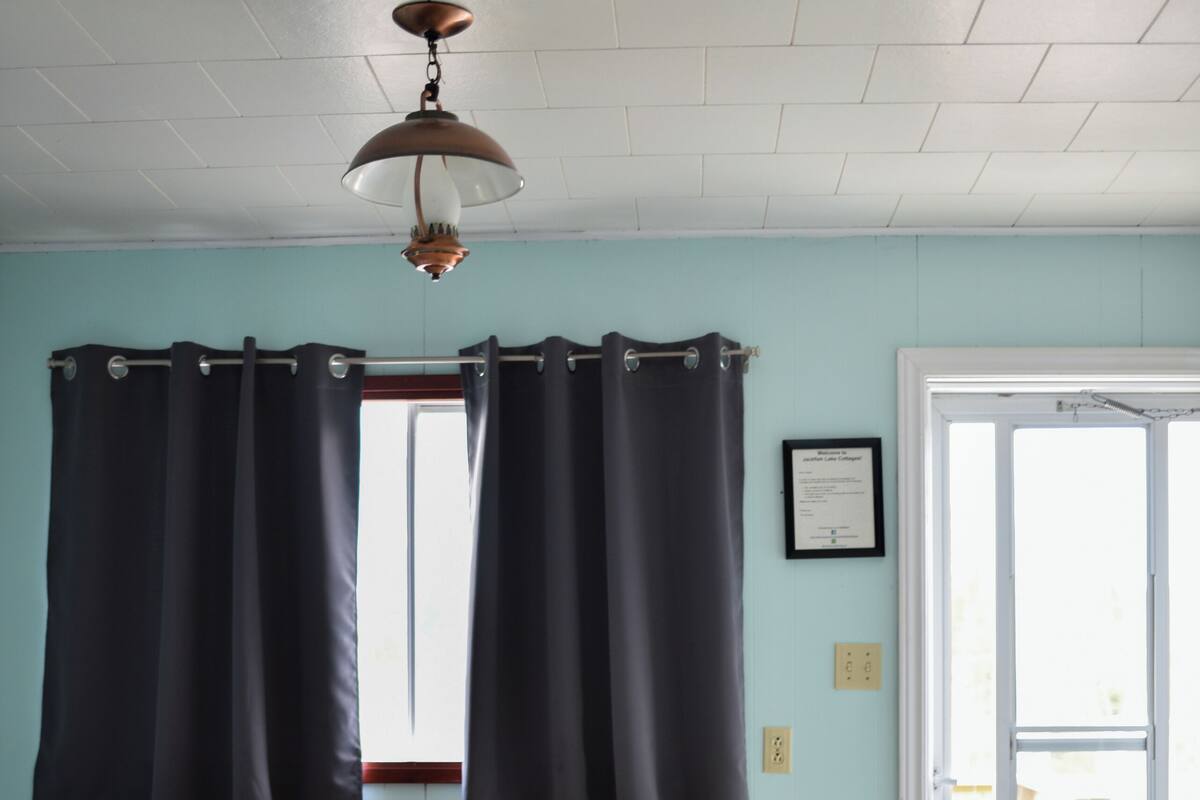 A room with a bed, a wooden table with chairs, a window with curtains, a door, and a ceiling fan.
