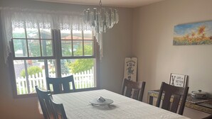 Un comedor con una mesa redonda, seis sillas, una araña, vistas a una cerca blanca y un cuadro en la pared.