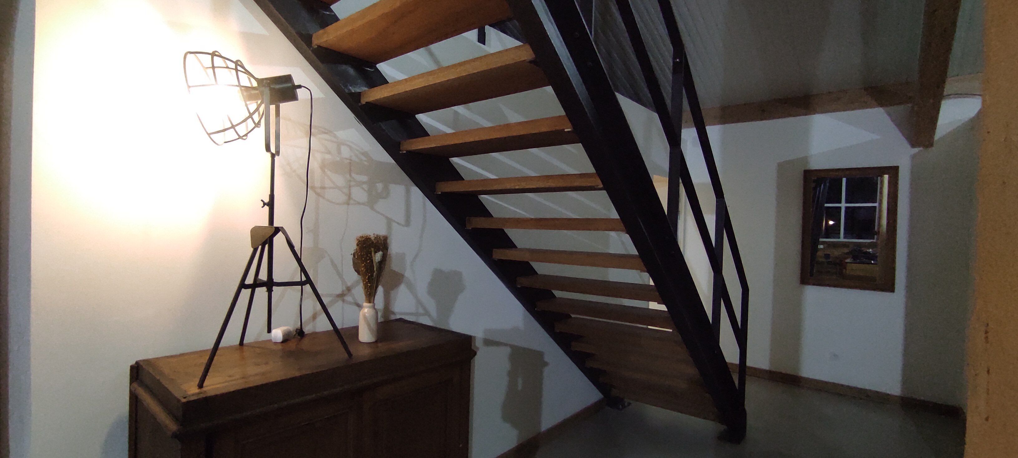 A staircase with wooden steps and black railings, a desk with a lamp, and a vase with dried flowers.