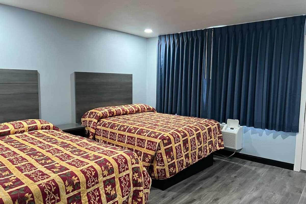 A hotel room with two beds, dark curtains, a ceiling light, and a wall-mounted air conditioning unit.