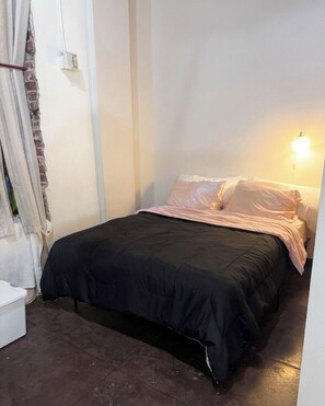 A small, single-bed room with a black bedspread, white pillows, a bedside lamp, and a white chair. - The Continental Royal Loft (Los Angeles)