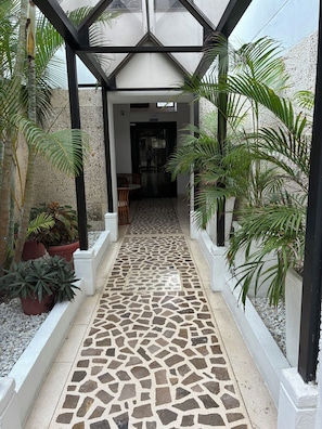 A covered walkway with a mosaic floor, flanked by potted plants and palm trees.