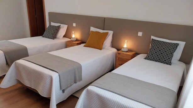Una camera d'albergo con tre letti singoli, ognuno con una coperta trapuntata bianca e una coperta grigia. Sono presenti comodini con lampade e una testiera in legno.