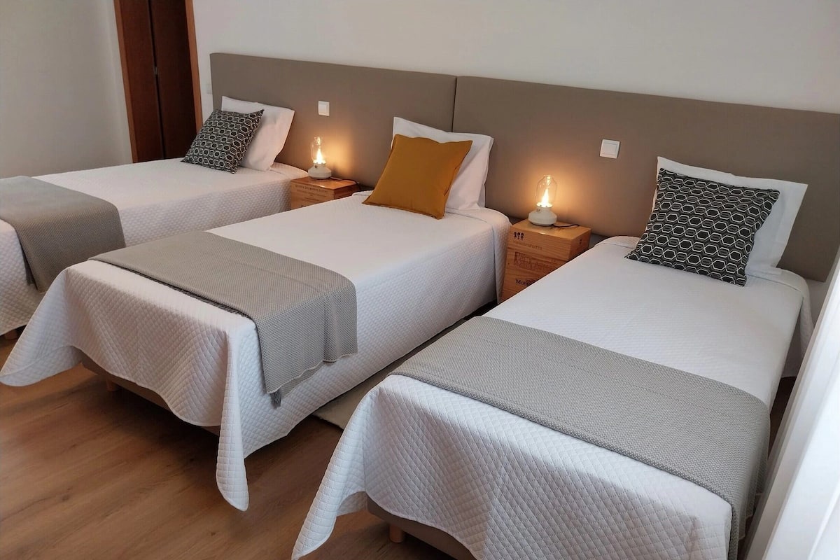 Ein Hotelzimmer mit drei Einzelbetten, jeweils mit weiß gestickter Bettdecke und grauer Tagesdecke. Nachttische mit Lampen und ein hölzernes Kopfteil sind vorhanden.
