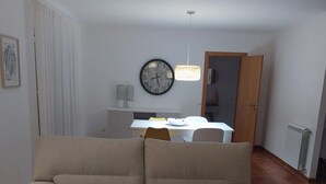 Un soggiorno con un divano color beige, un tavolo da pranzo con sedie, una sveglia e una lampada a sospensione.