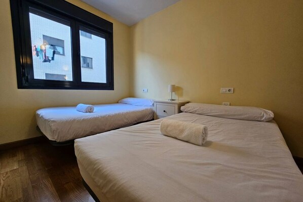 A room with two single beds, a nightstand, a lamp, and a window with a view of a building.