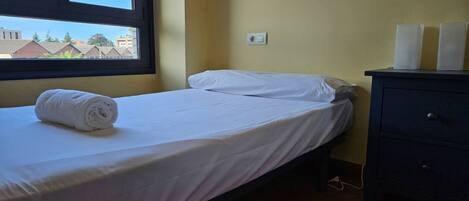 A single bed with white linens, a window with a view of buildings, a wall socket, and a dark-colored dresser.