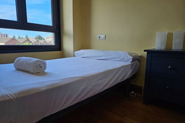 A single bed with white linens, a window with a view of buildings, a wall socket, and a dark-colored dresser.