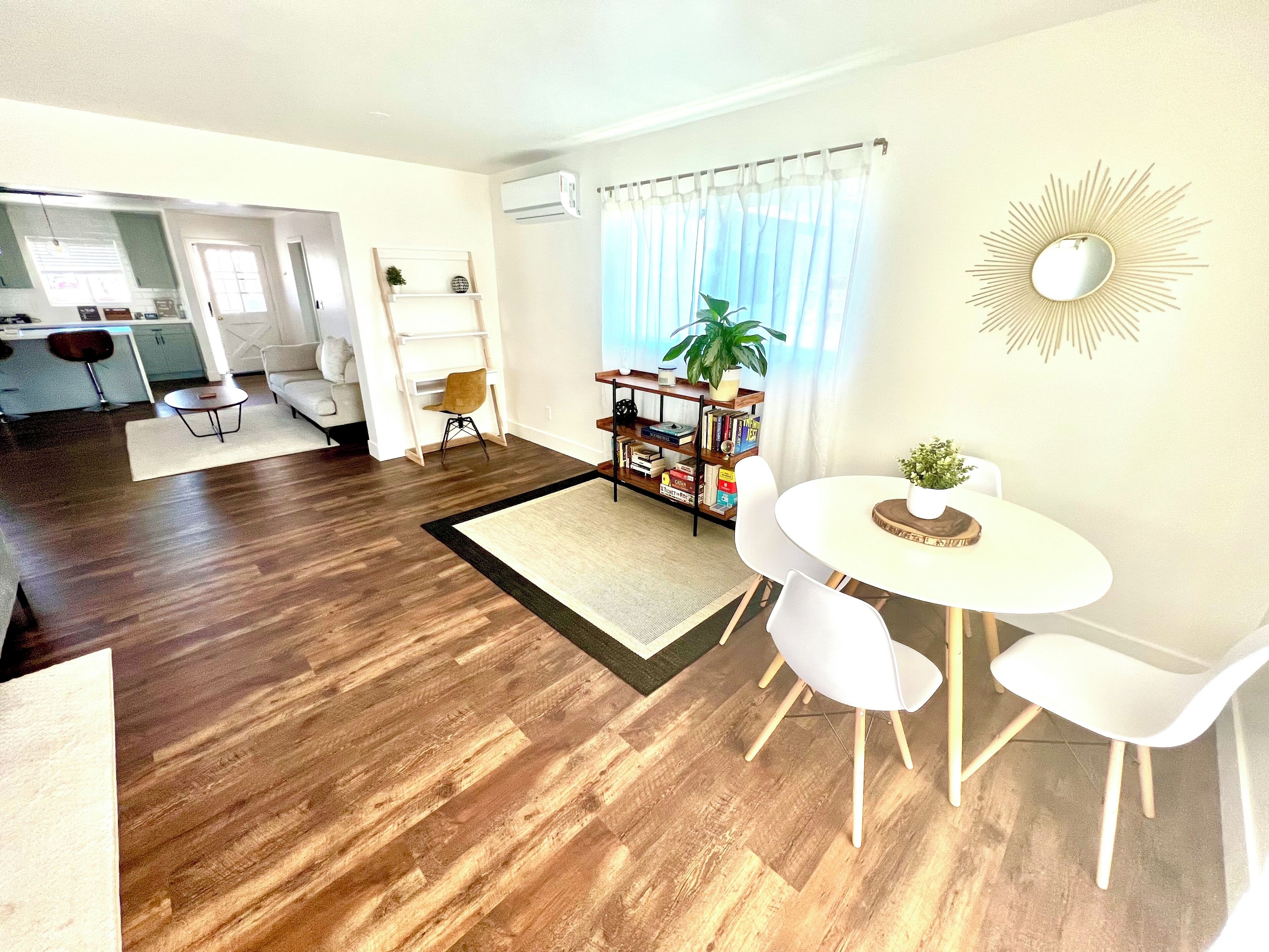 A modern living room with a dining area, wooden flooring, white furniture, and a bookshelf with books and plants.