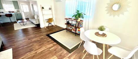 A modern living room with a dining area, wooden flooring, white furniture, and a bookshelf with books and plants.