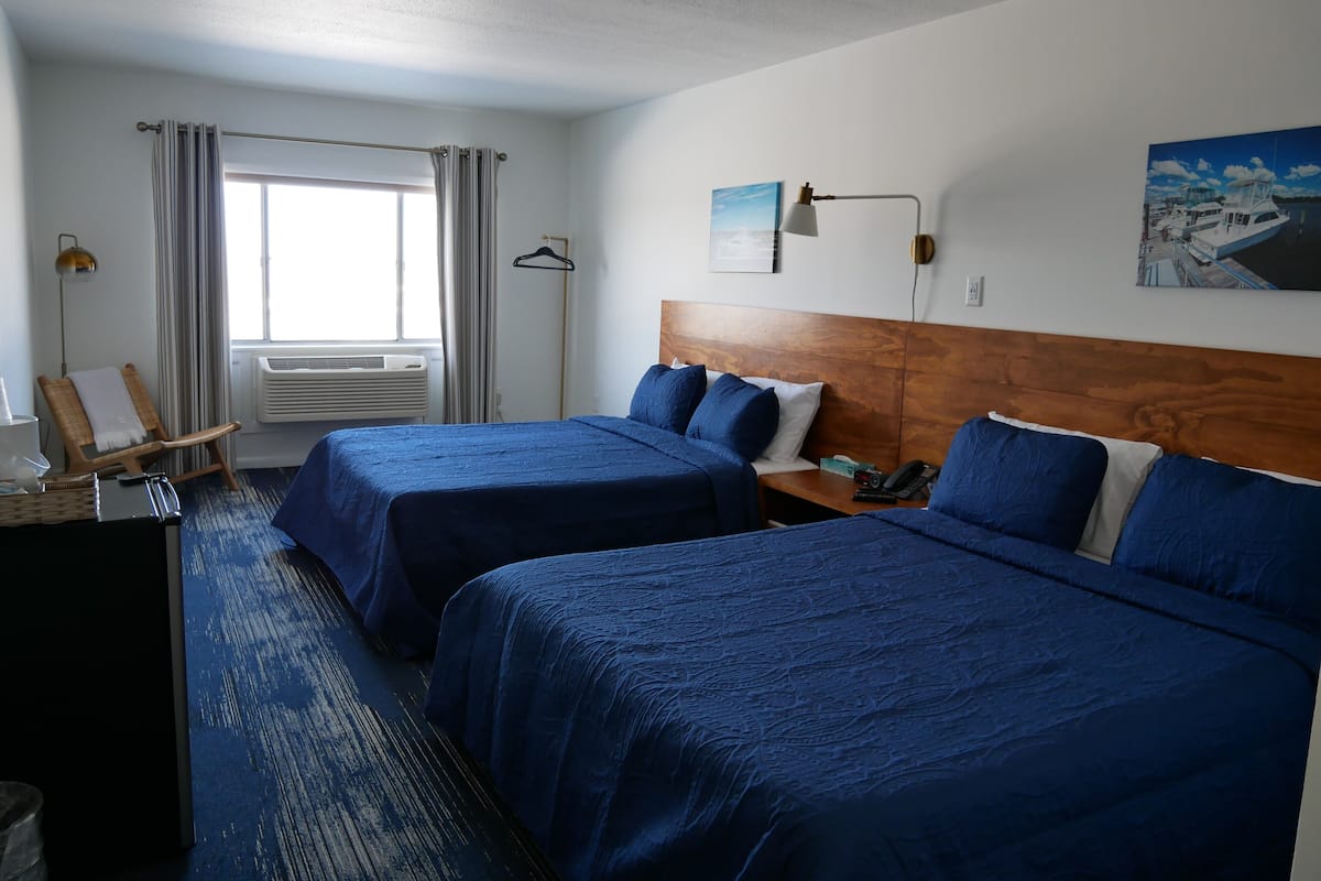 A hotel room with two beds, a wooden headboard, a nightstand, a lamp, a painting, and a window with curtains.