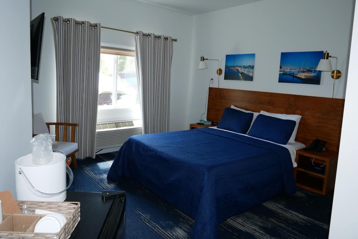 A hotel room with a bed, a chair, a table with a basket, a lamp, and two paintings on the wall.