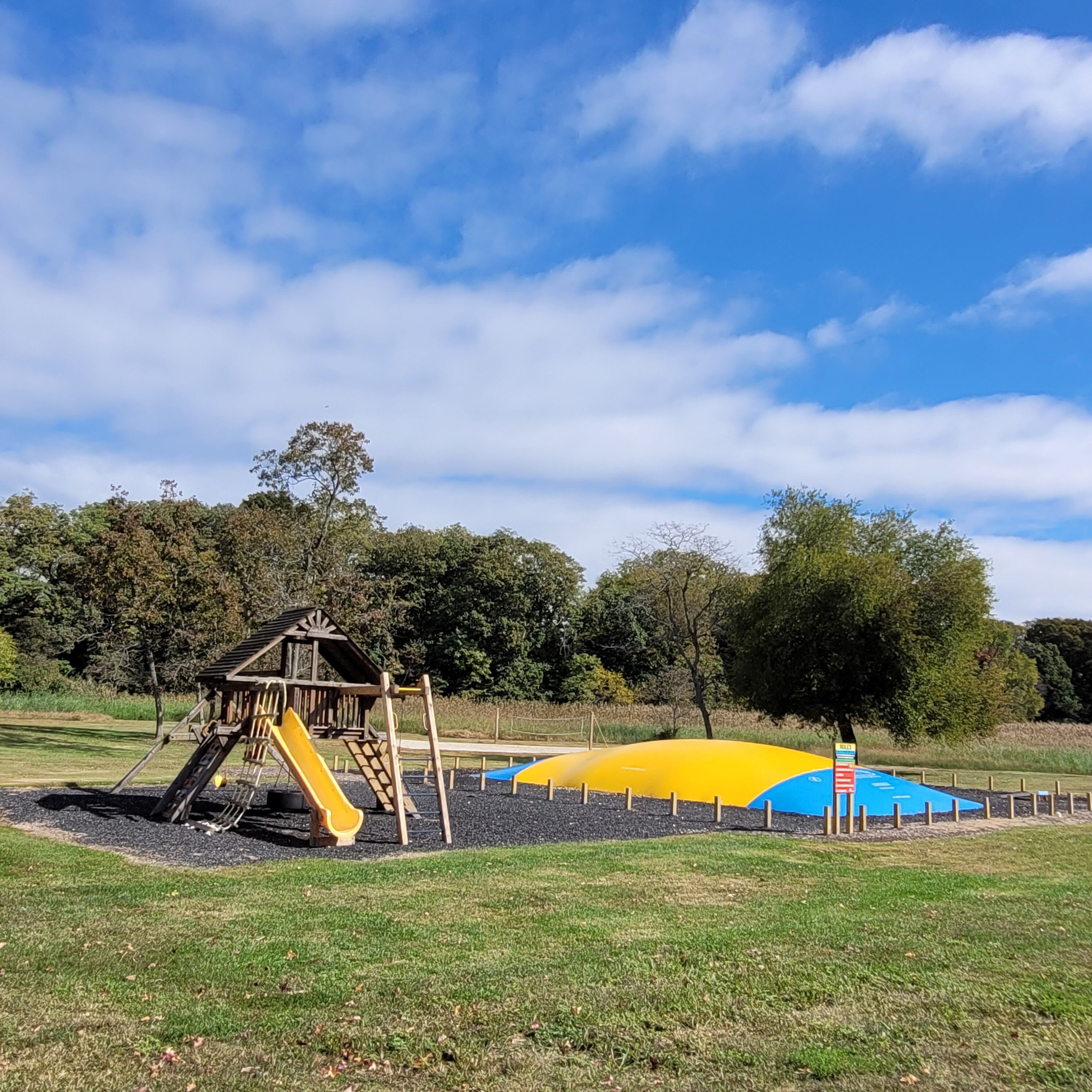 Un terrain de jeux avec un toboggan en bois et une structure d’escalade, entouré d’herbe et d’arbres, sous un ciel dégagé.