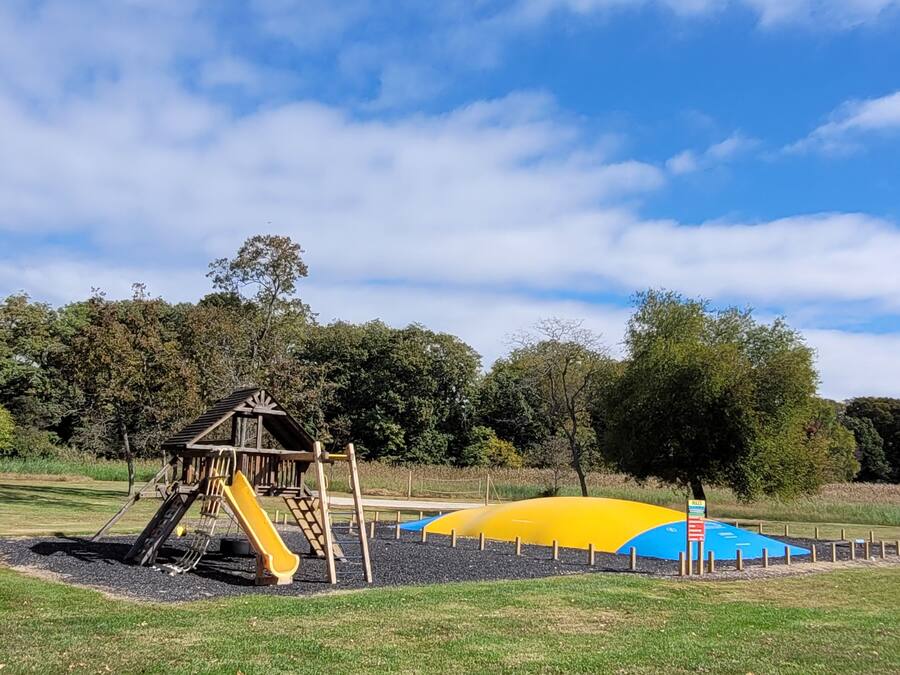 Un terrain de jeux avec un toboggan en bois et une structure d’escalade, entouré d’herbe et d’arbres, sous un ciel dégagé.