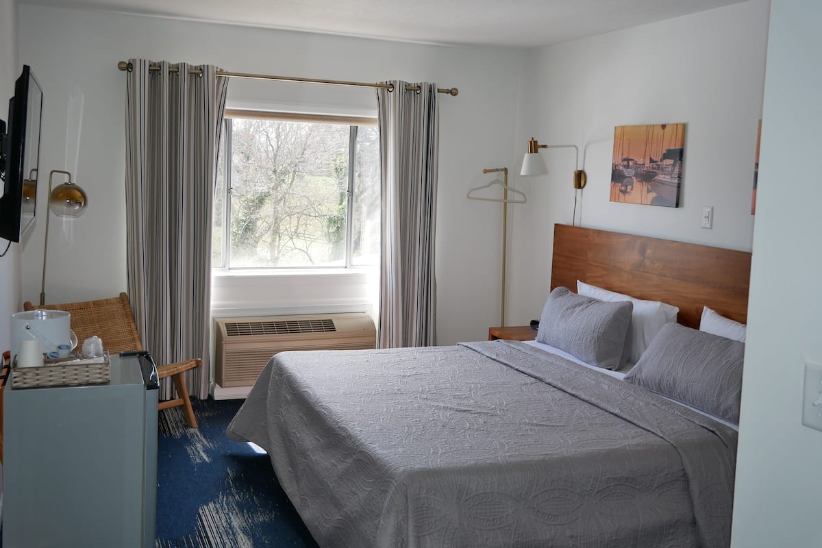 A hotel room with a bed, a desk, a chair, a window with curtains, a wall-mounted TV, a painting, and a floor lamp.