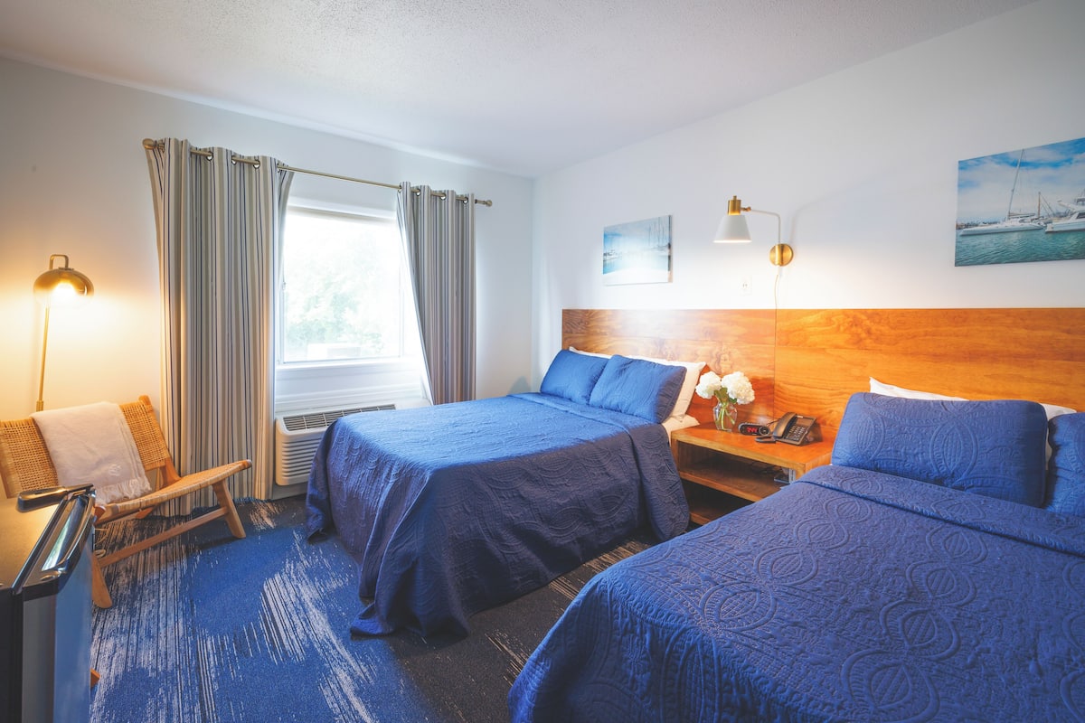 A hotel room with two beds, a wooden headboard, a desk with a lamp, a chair, and a window with curtains.