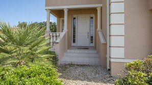 Una casa con fachada color beige, columnas blancas, una puerta blanca y un pequeño porche con escalones que conducen a la puerta.
