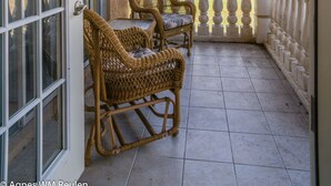 Un balcón con dos sillas de mimbre, un ventilador de techo y vistas a casas y palmeras.