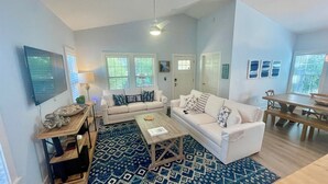 A spacious living room with two white sofas, a coffee table, and a dining table. There are large windows with blinds, a ceiling fan, and a flat-screen TV mounted on the wall. - Family Friendly | Walk to Beach & Dining | Firepit (Santa Rosa Beach)