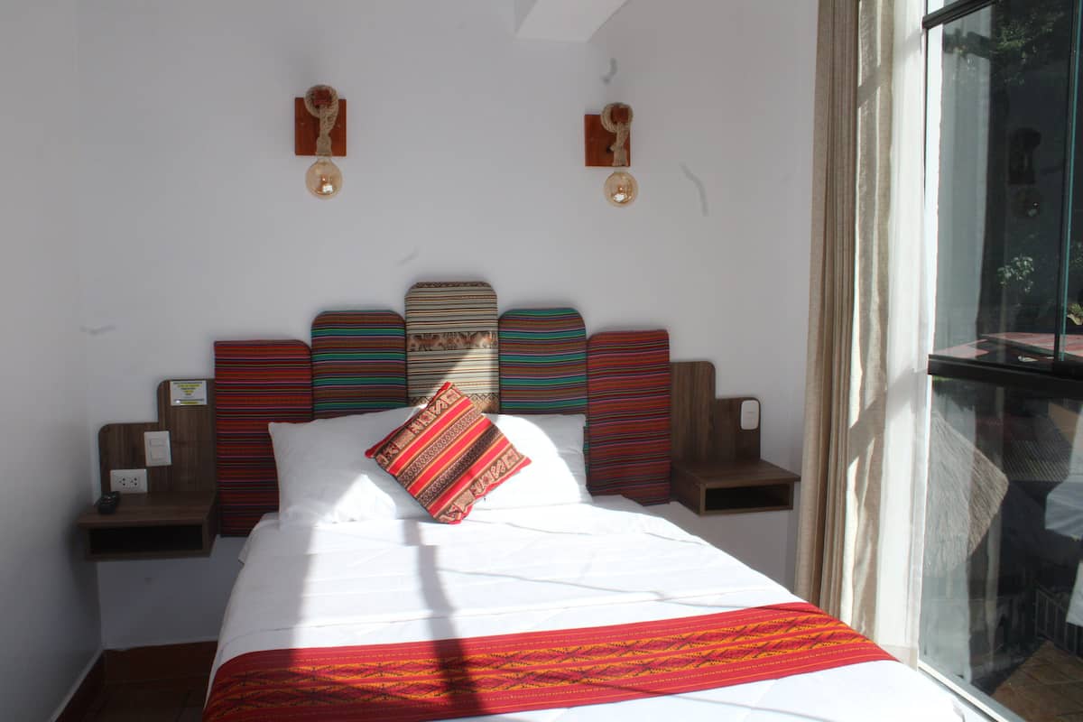 A neatly made bed with a red and white bedspread, a striped pillow, and a headboard with multiple panels. There are bedside tables with lamps and a window with a view of the outside.