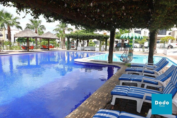 Una piscina con sillas de descanso y sombrillas, rodeada de palmeras y un área de estar cubierta.