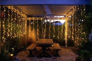 An outdoor patio with a wooden table and benches, surrounded by greenery and string lights. - Newly Furnished, Entire Home, Pool House (Los Angeles)