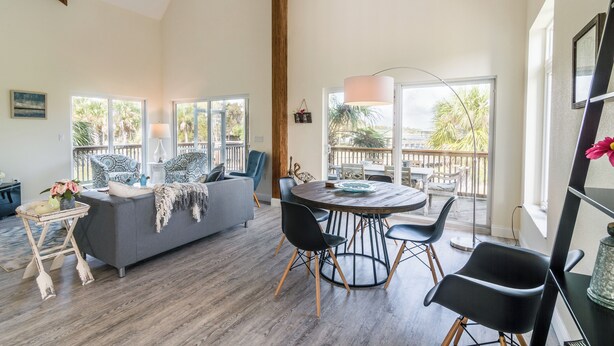 A spacious living area with a grey sofa, a round wooden table with chairs, and large windows offering a view of palm trees.
