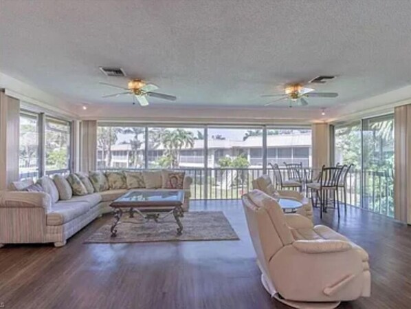 Relax in this spacious living area with a high ceiling, ceiling fans, and a large window offering a view of palm trees and a building in the distance.